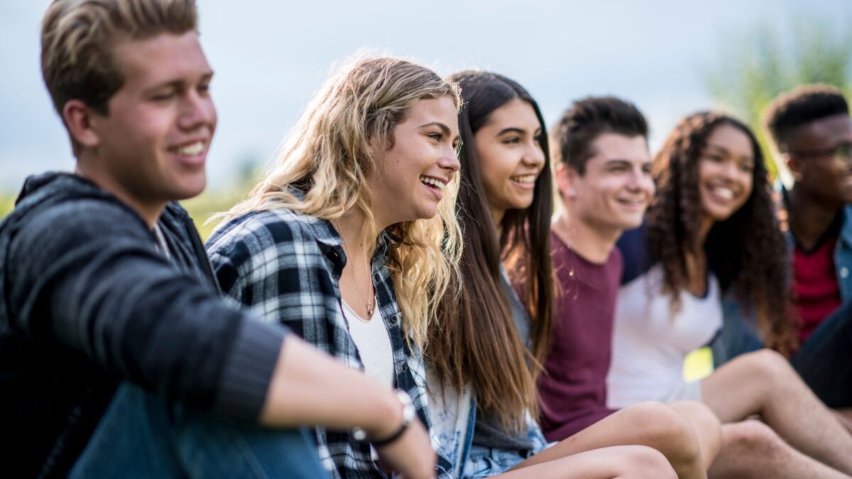 Six teenagers, three boys and three girls of various races sit together looking happy. 