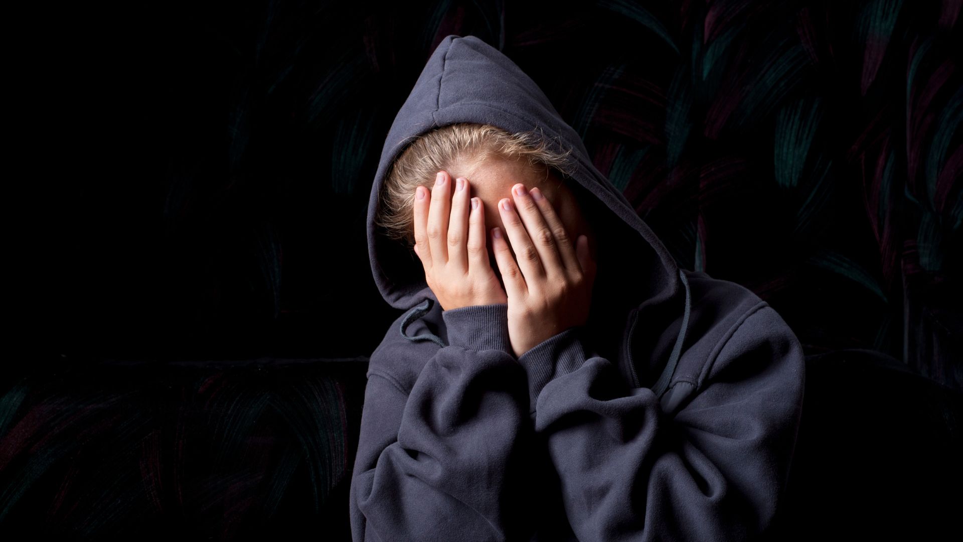 Teenage girl wearing a blue hoodie holds her face in her hands, looking sad and upset.