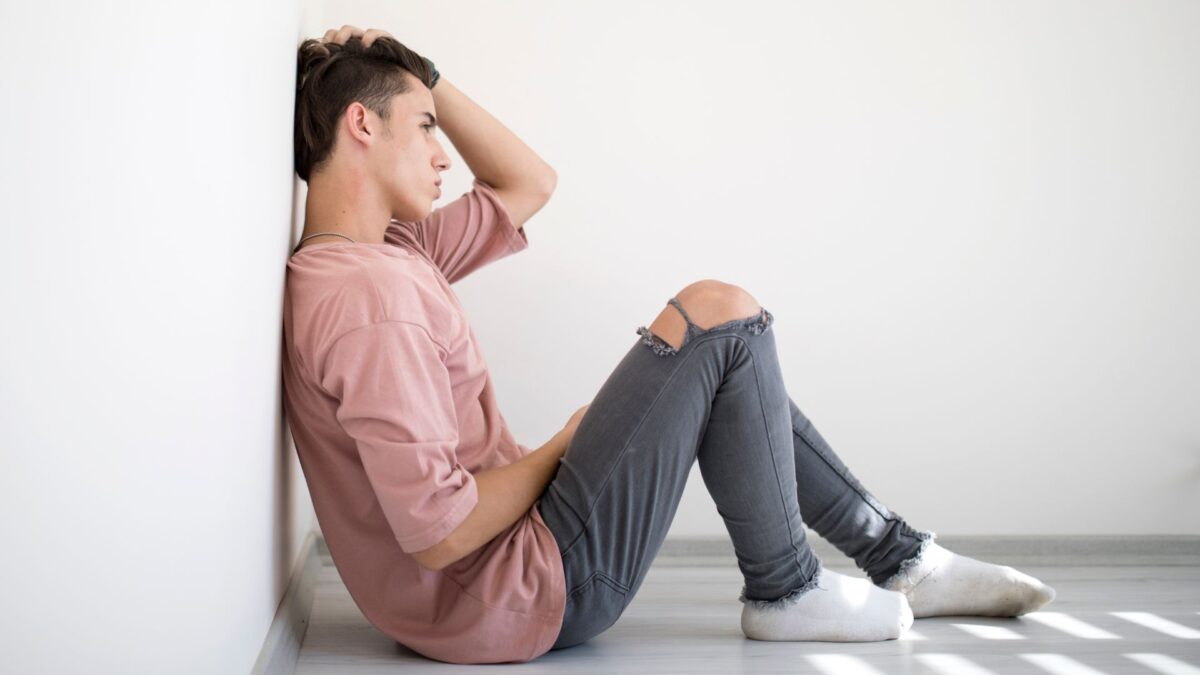 An older teenage boy wearing a sweatshirt, jeans with holes in the knees and socks sits on the floor of a room, leaning against a white wall, looking sad. 