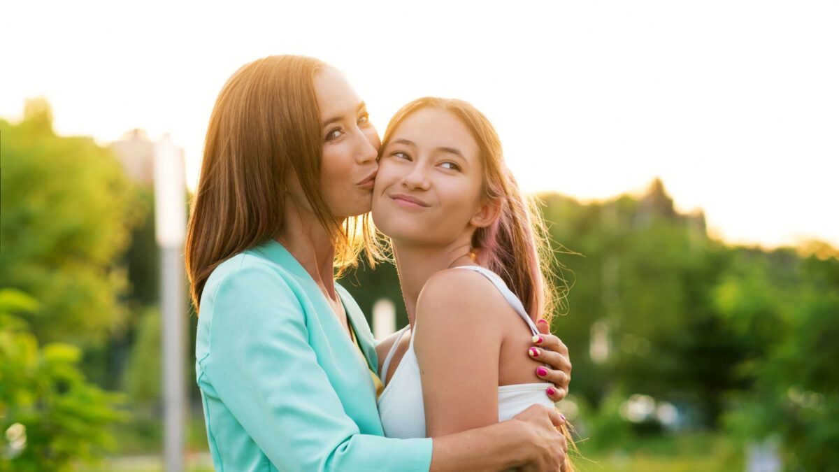 A mom in a blue blazer kisses her daughter, who wears a white dress. They appear to be standing in front of a setting sun, and a row of trees. 