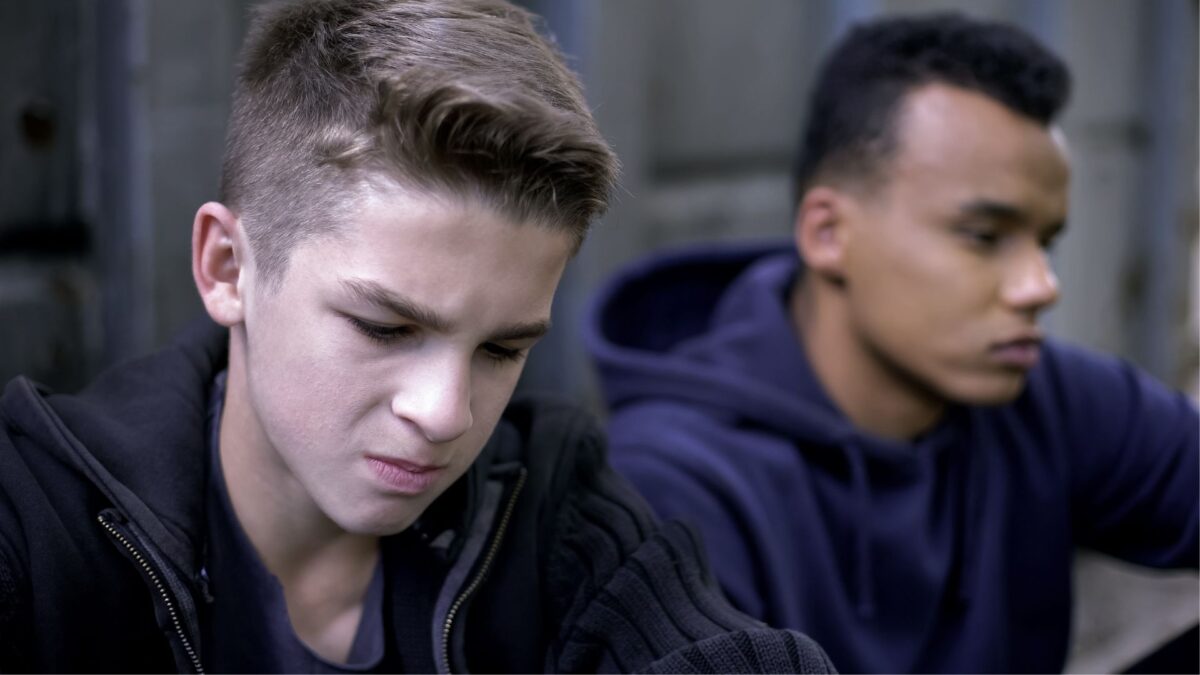 A teen boy sits next to another teen, looking sad and angry.