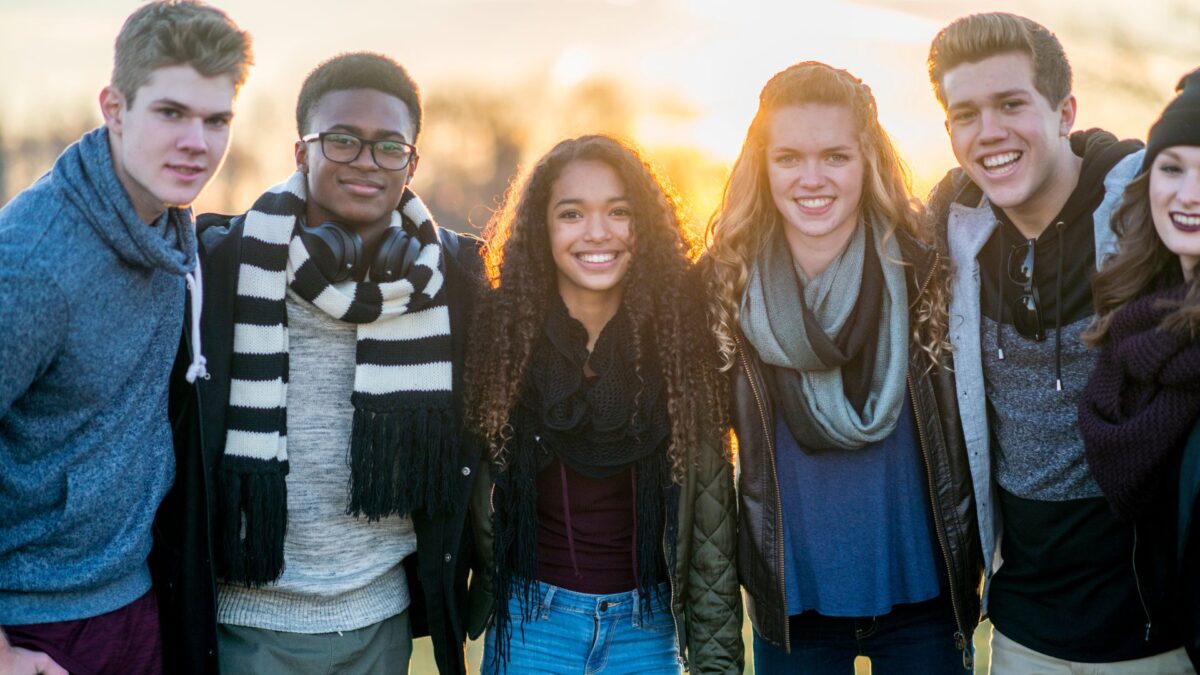 Six teens wearing fall clothing smile in front of a sunset in a park-like setting. 