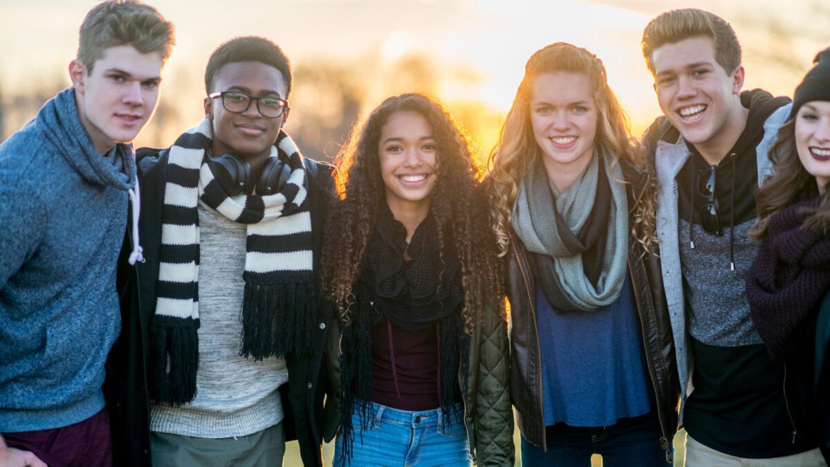 Six teens wearing fall clothing smile in front of a sunset in a park-like setting. 