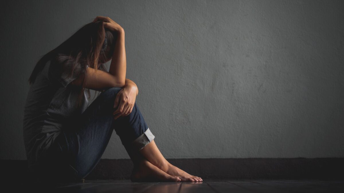 A teenager girl sits alone in a dark space, holding her head close to her knees, looking sad. 