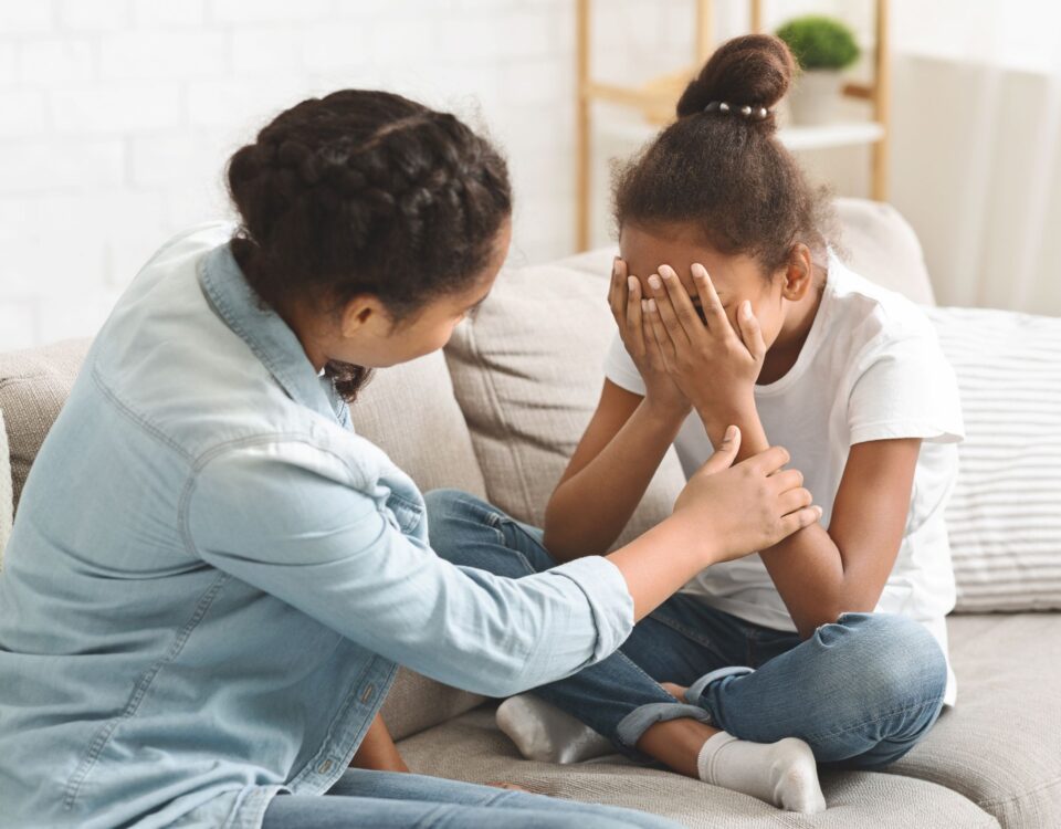 A mother tries to comfort her crying daughter on the couch.