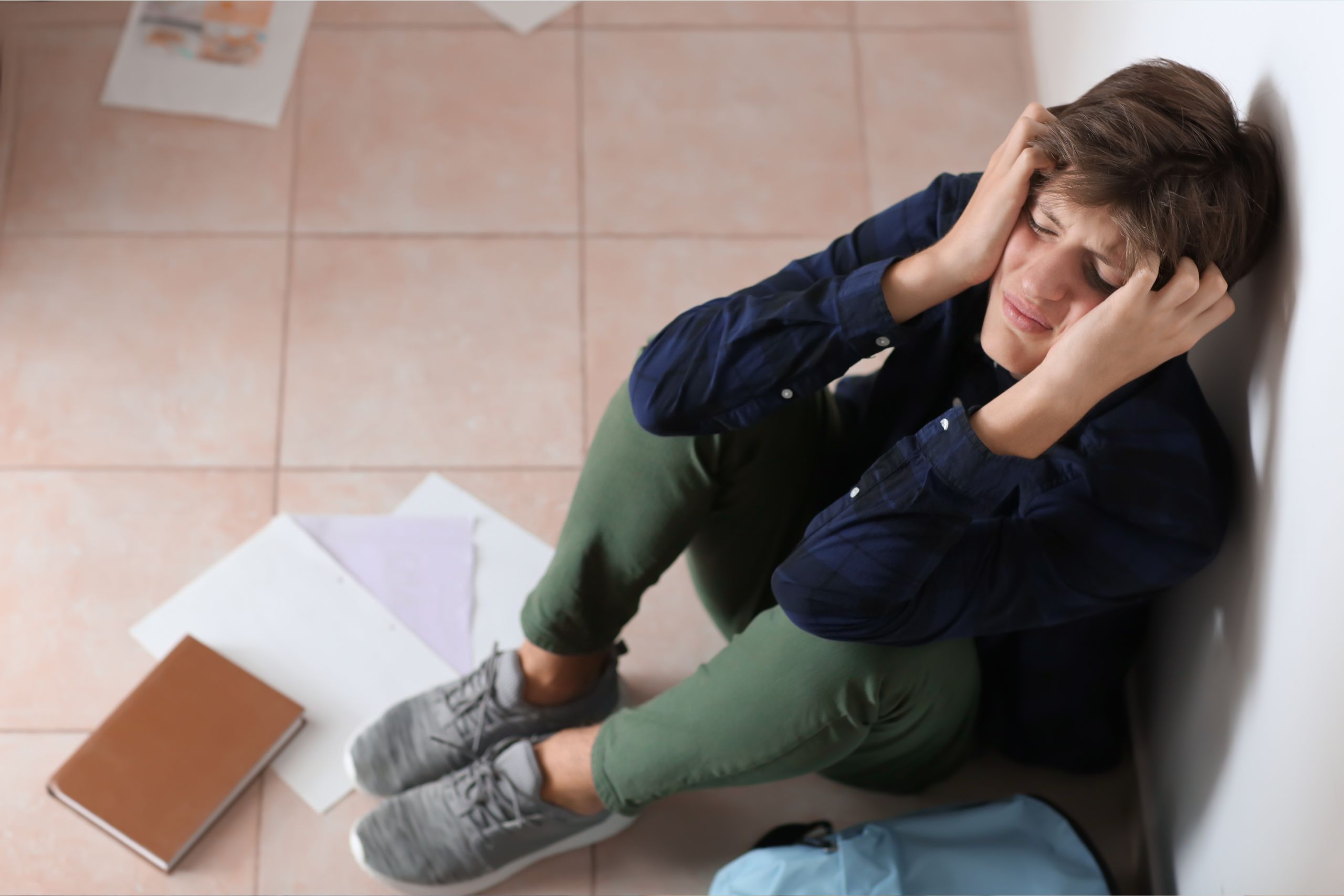 A teenage boy sits on the floor, with a book and papers dropped near him. He is holding his head with both hands, as if he is sad or scared.