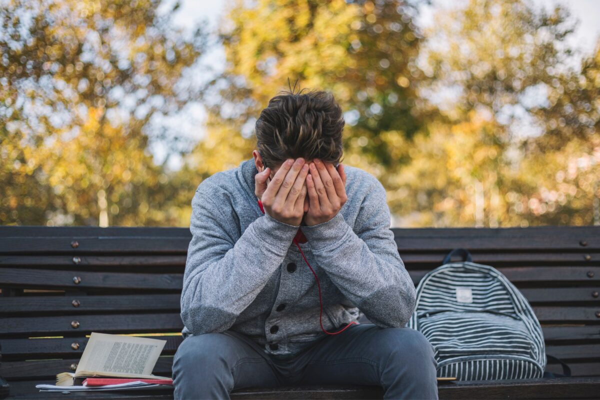 A teenage boy sits on a park bench, covering his eyes with his hands. 