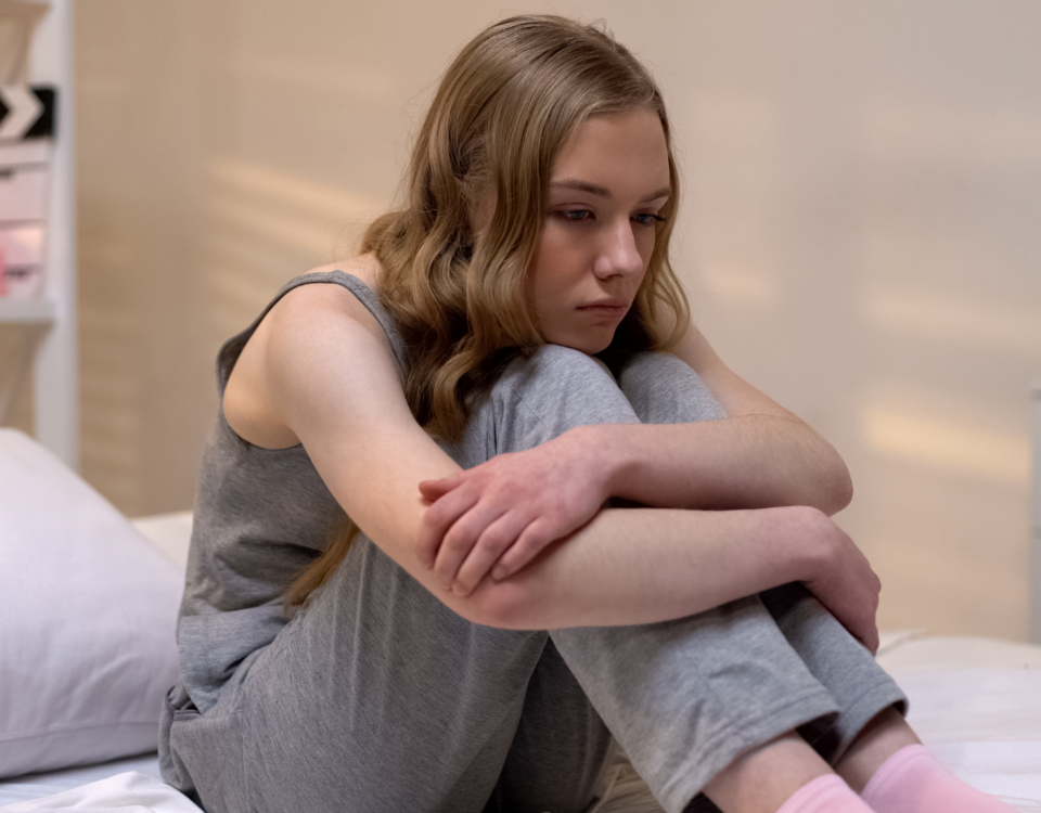 teen girl on a bed with her chin resting on her knees and her arms crossed over her shins.