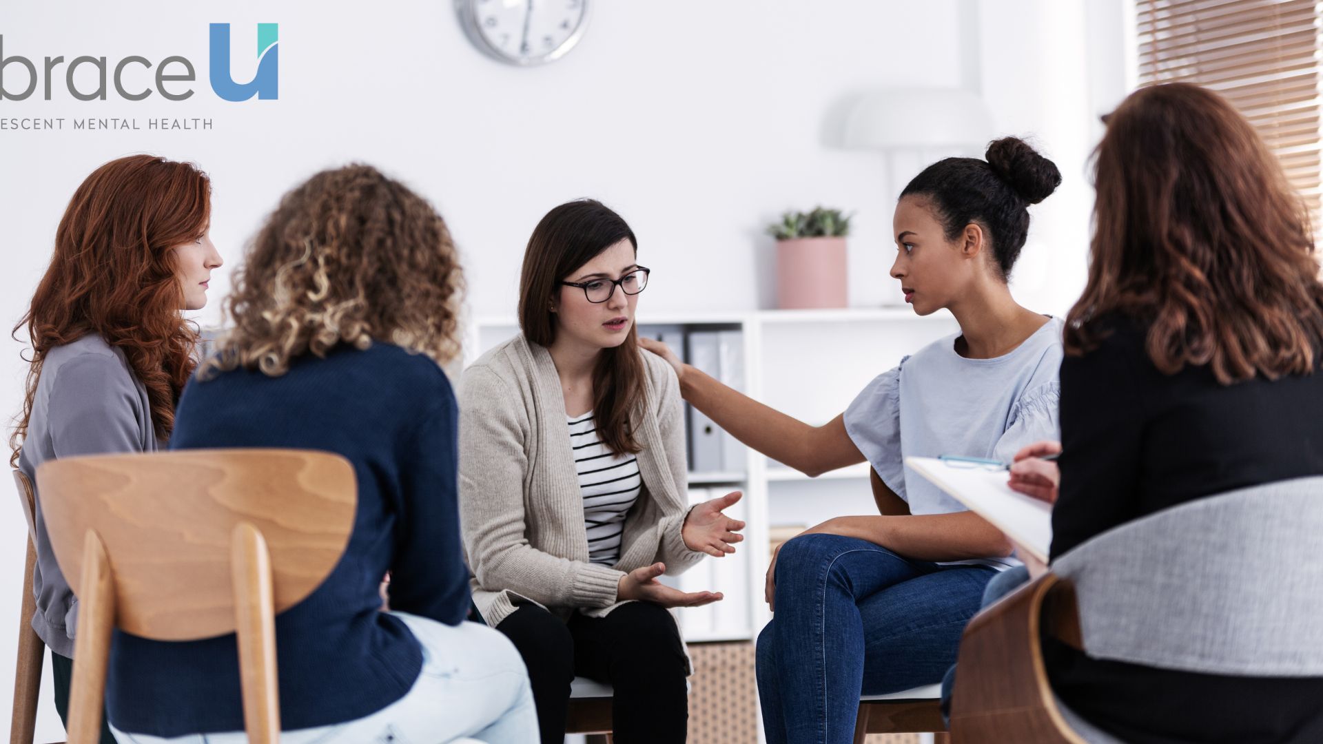 A group of young women hold a therapy session in a white room. One girl is touching the shoulder of another to comfort her.