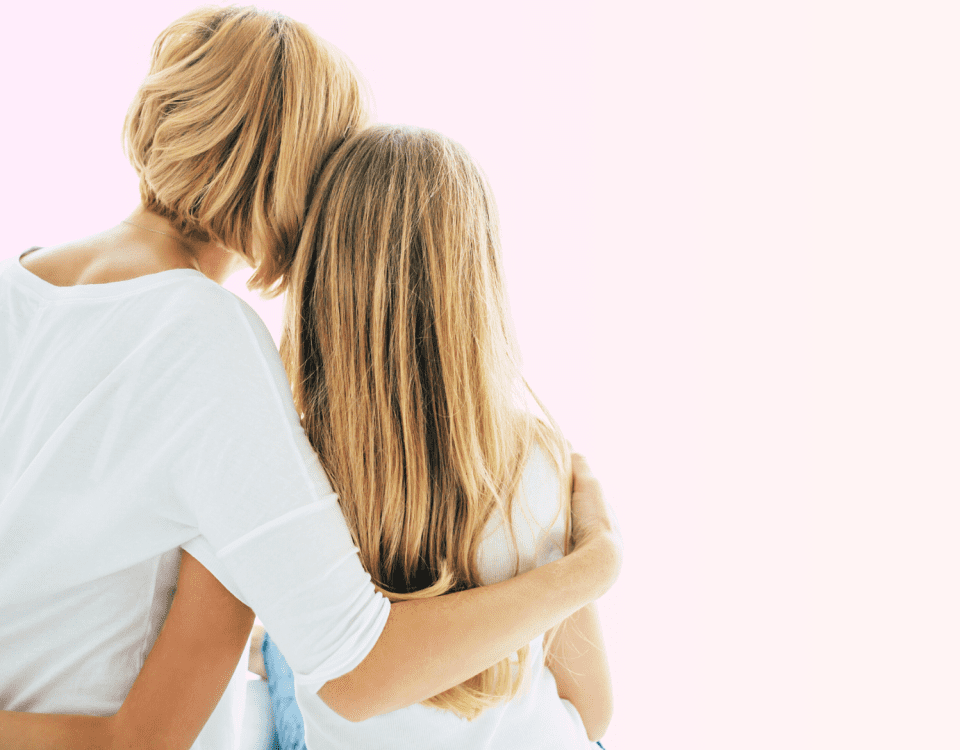 A mother and daughter sit with their backs to the camera and their arms around each other. The mother has short blonde hair, the daughter has long blonde hair. Both are wearing white shirts.