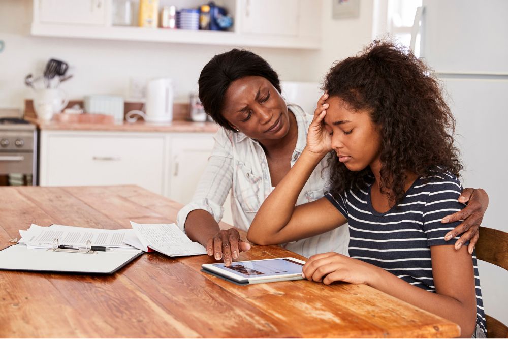 Teen depression impacts more than 3% of adolescents each year. In this picture a mother puts her arm around her teenage daughter as they sit at a kitchen table.