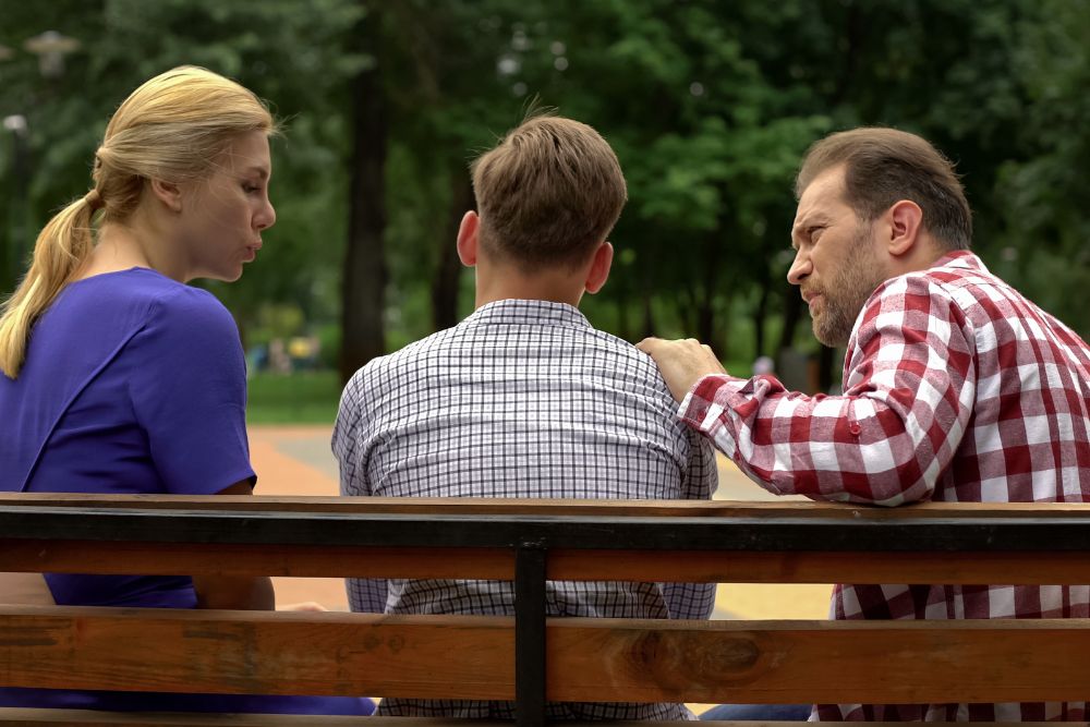 When should parents consider more intensive treatment for a child with mental health issues? In this photo a mom and dad sit on other side of a teenage boy on a park bench. They are having a serious conversation about mental health.