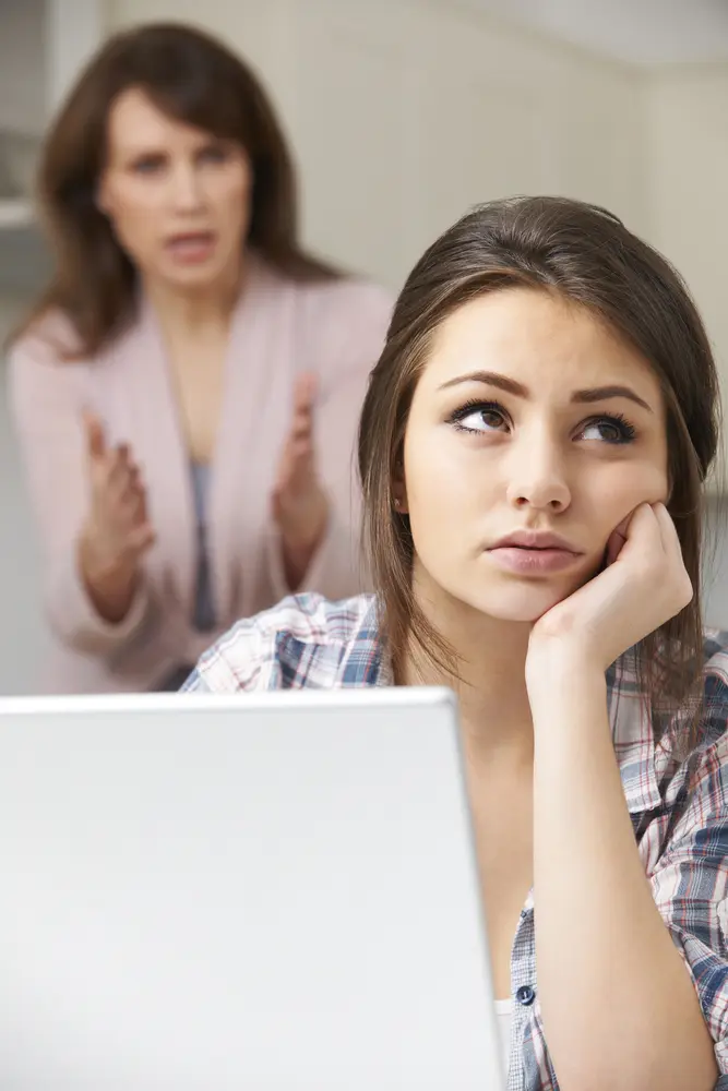 Teen seated at her computer and rolling her eyes and her mother in the background.