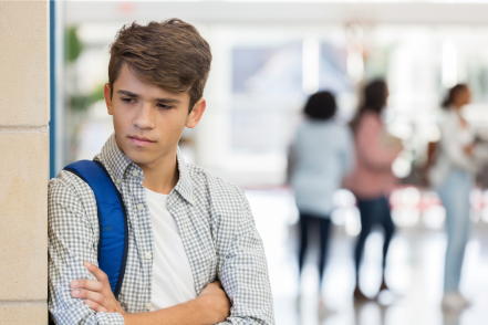 A teenage boy wearing a backpack leans against a wall in school looking sad as other students walk away behind him.