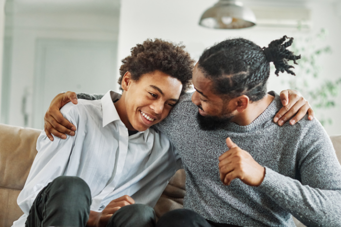 A father is talking to his son on a couch, with his arm wrapped around him. Both appear to be smiling.
