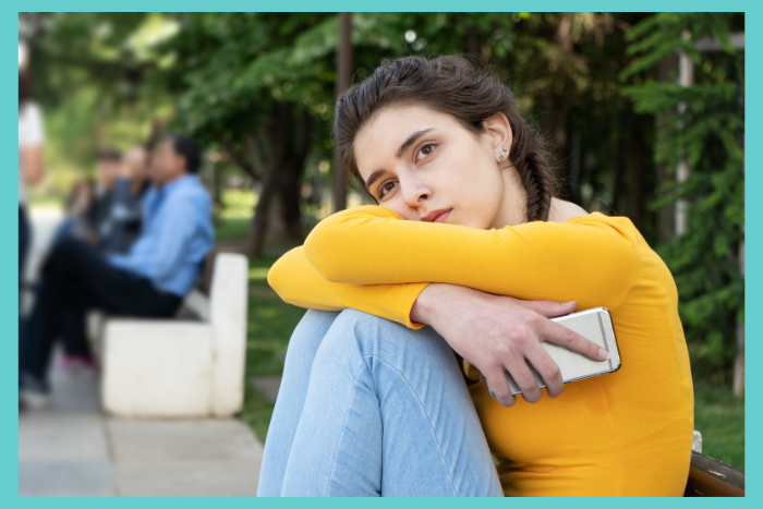A girl sits on a bench alone holding a phone and looking sad to express teen loneliness.