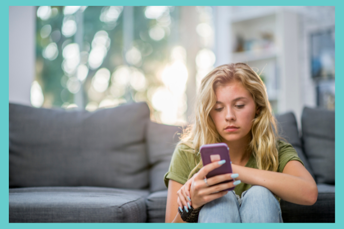 Blonde teen girl sits in front of a couch looking at her cell phone.