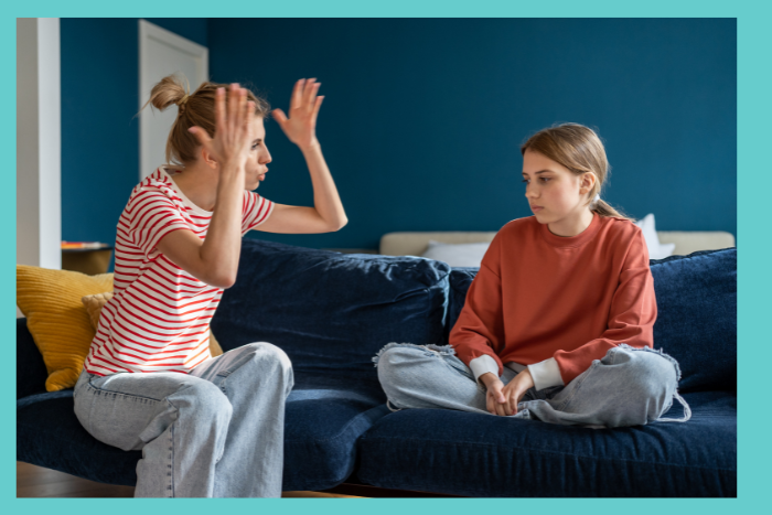 A mother raises her hands in anger while having an argument with a teenage girl who sits cross legged on a couch.