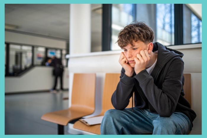 A teenage boy sits in an emergency room waiting area. He appears sad or upset. He is wearing a hoodie and holds his head in his hands.