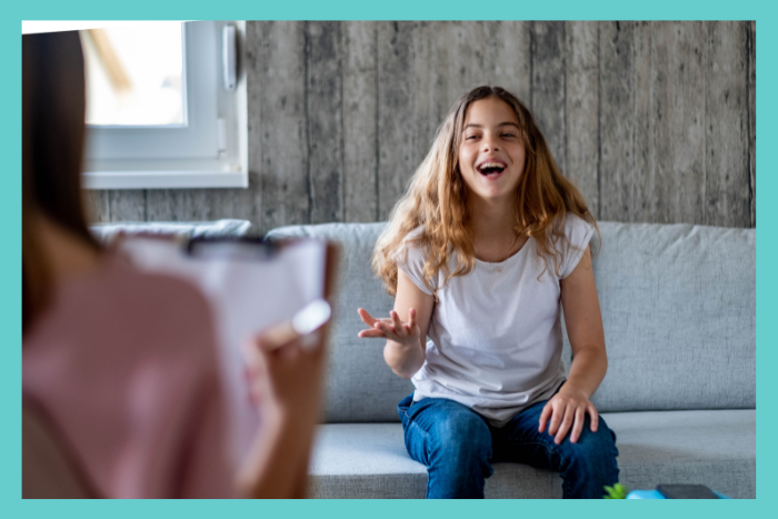 A young teenager girl sits on a couch in a therapist's office. She is laughing and appears to be having fun.