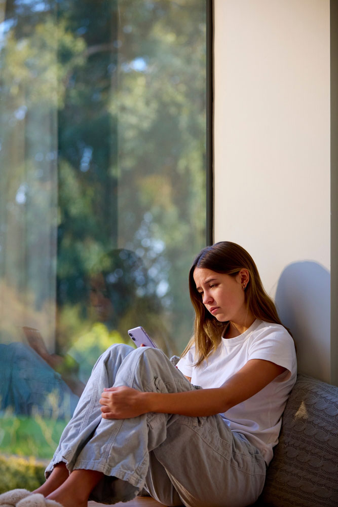 Depressed teen sitting on windowsill looking at her phone