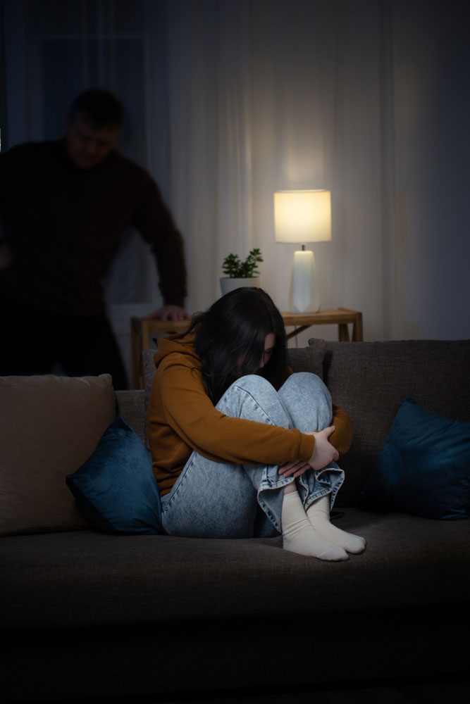 Depressed teen sitting on couch in dark with holding legs to body with parent in the background