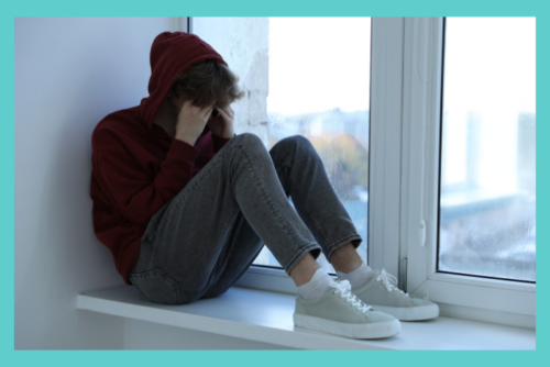 depressed teen boy sitting near a windowsill covering their face; depression