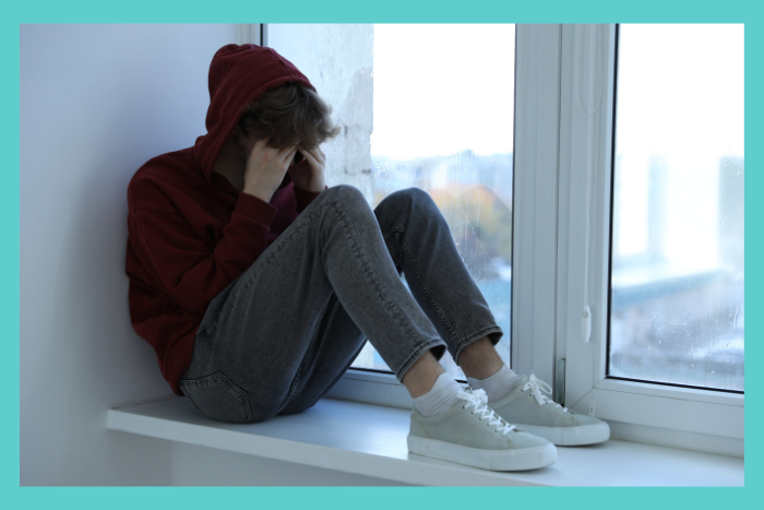 depressed teen boy sitting near a windowsill covering their face; depression